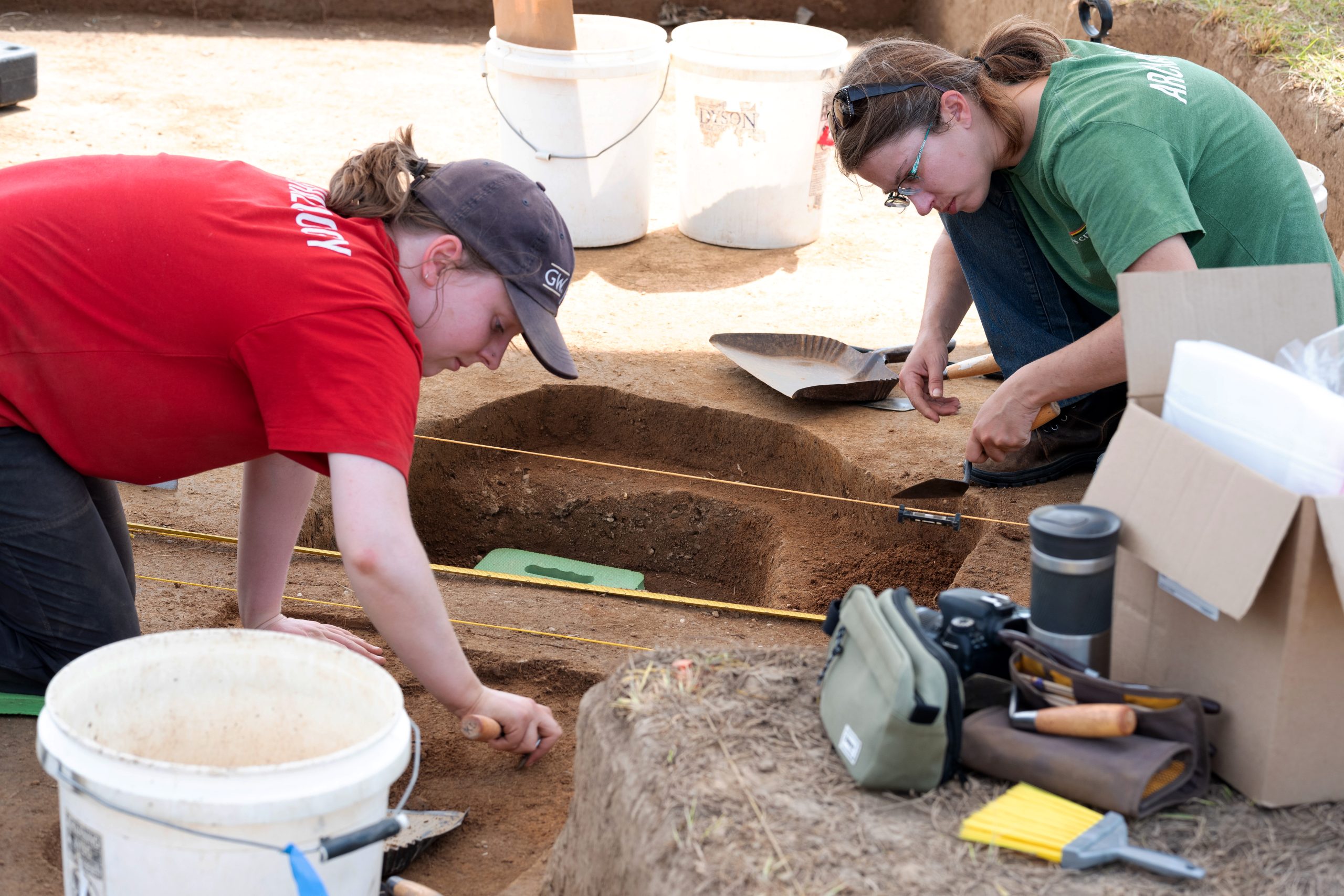 Two people are working at an archaeological dig site. One has a trowel in hand and is digging in the soil.