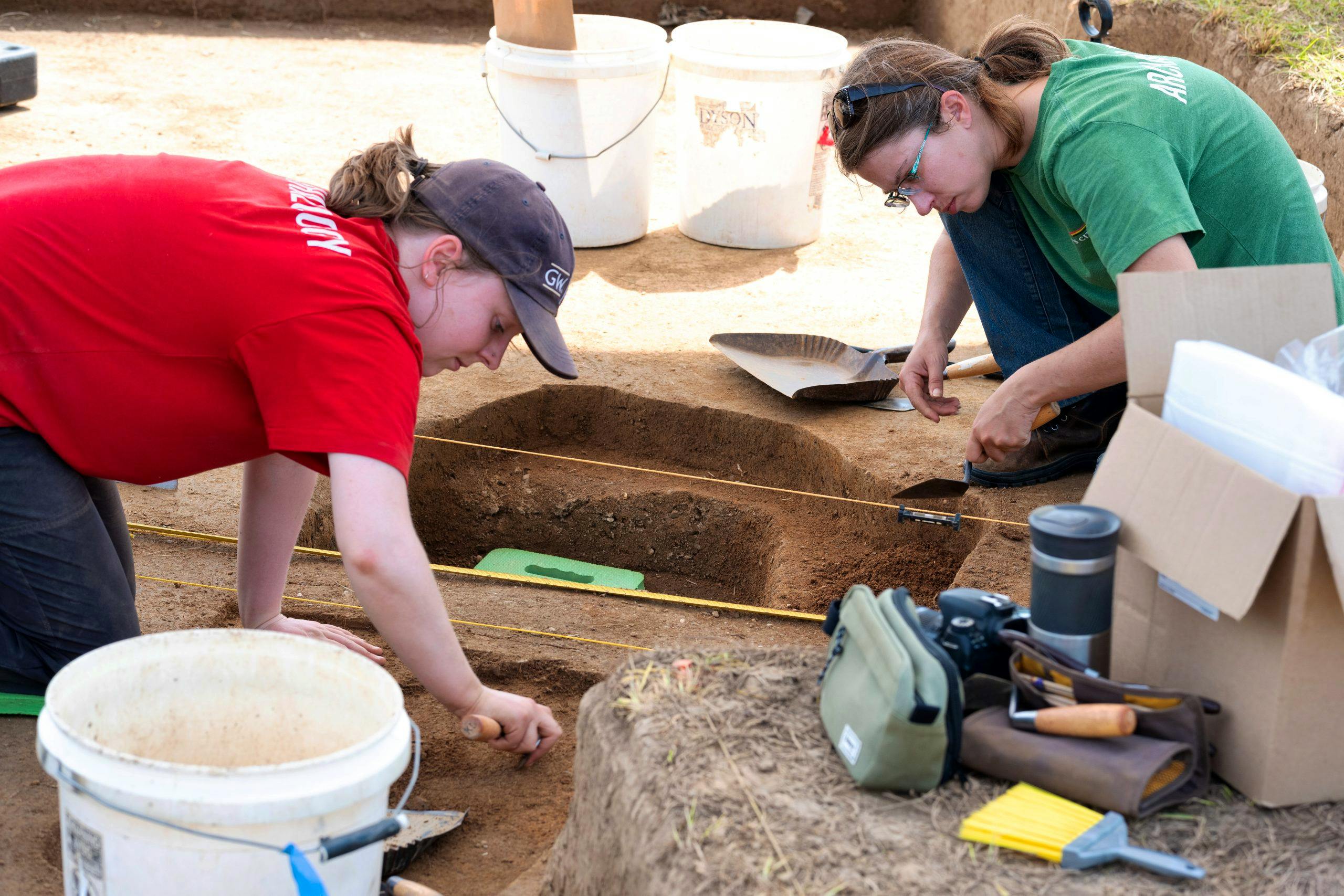 Two people are working at an archaeological dig site. One has a trowel in hand and is digging in the soil.