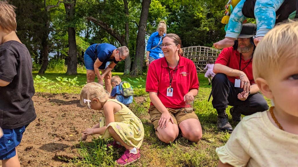 An HSMC staff member is sitting in a garden, smiling. They are sitting in soil,with their left hand closed. Next to them is a toddler kneeling with their back facing the staff member. The child is planting a seed in the soil. Behind both of them is a caregiver helping their child plant in the soil.