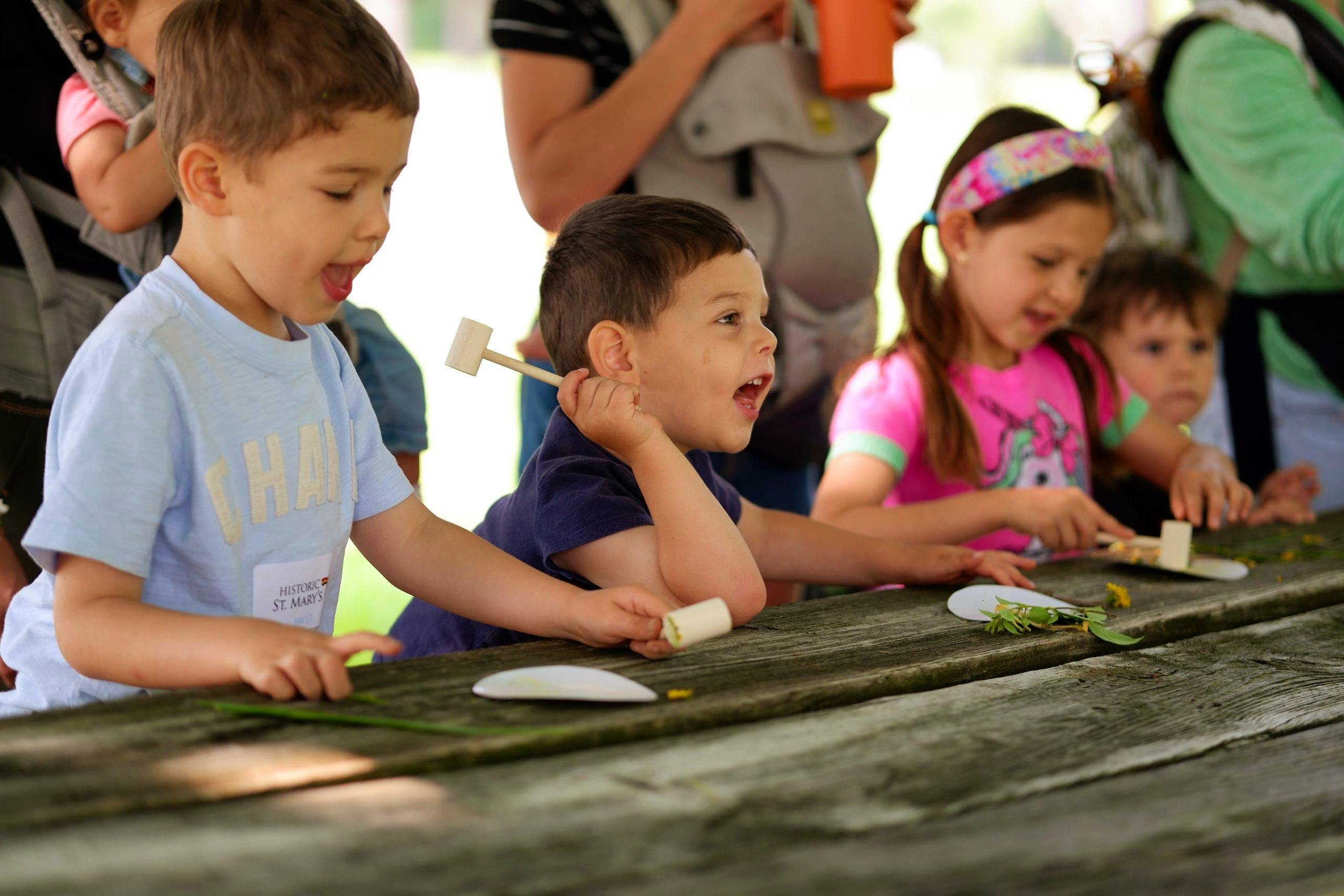 Three children are sitting outside at a wooden picnic table. They are participating in the Little Explorers preschool program. They each have a little wooden mallet in their hand and a small paper plate in front of them on the table. Next to the plate is a small gathering of leaves and flowers. They each have an excited look on their faces.